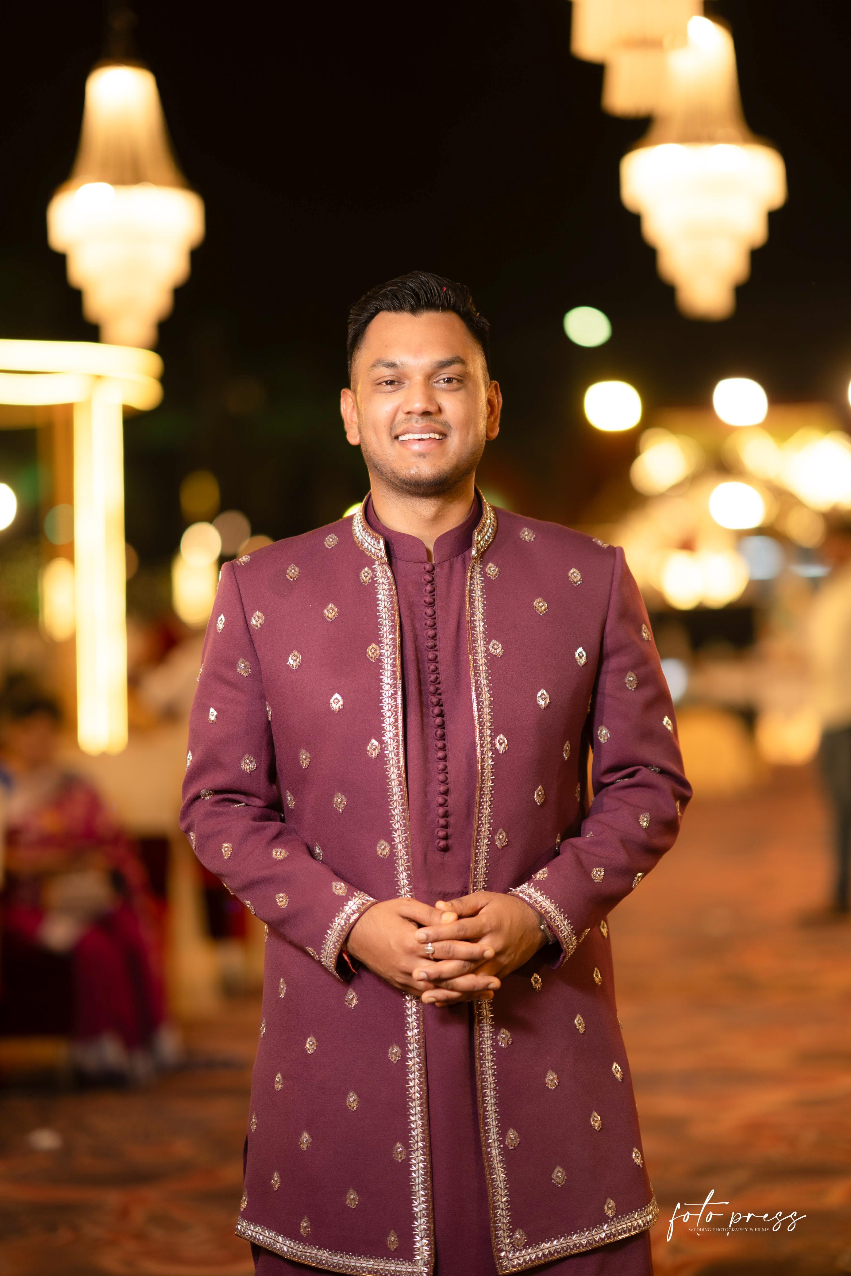 Man wearing a maroon sherwani with gold patterns standing against a blurred background with lights.